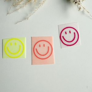 May include: Three square stickers with smiley faces. One is light green with a yellow smiley face, another is peach with a red-orange smiley face, and the third is white with a magenta smiley face.