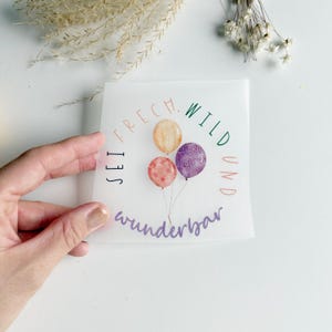Könnte beinhalten: Ein quadratischer Aufkleber mit dem deutschen Text "SEI FRECH. WILD UND wunderbar" in kreisförmiger Anordnung. Der Aufkleber zeigt drei Aquarellballons in Orange, Rosa und Lila. Der Aufkleber wird von einer Hand gehalten.