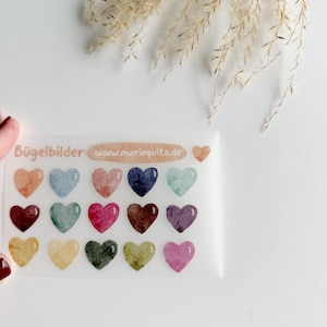 Könnte beinhalten: Ein Bogen mit aufbügelbaren herzförmigen Aufklebern in verschiedenen Farben, darunter Orange, Blau, Rosa und Grün. Der Text "Bügelbilder" und "www.mariequita.de" sind auf dem Bogen aufgedruckt.