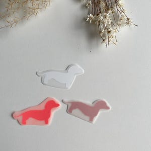 Könnte beinhalten: Drei gestanzte Aufkleber in Form von Dackeln. Ein Aufkleber ist einfarbig weiß, einer ist in leuchtendem Neonpink und einer in einem gedeckten Altrosa gehalten. Jeder Aufkleber hat einen durchscheinenden Rand um die Hunde-Silhouette.