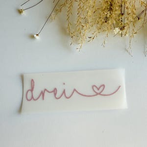 Könnte beinhalten: Ein rechteckiger weißer Aufkleber mit dem Wort "drei" in altrosa Schreibschrift, das in einem kleinen Herz endet. Die Schrift ist fließend und durchgehend gestaltet und für die Anbringung auf einer flachen Oberfläche vorgesehen.