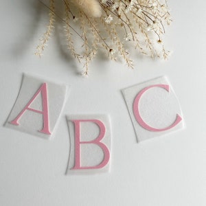 Könnte beinhalten: Drei einzelne Aufbügler mit den Buchstaben A, B und C in einem zarten Rosaton. Jeder Buchstabe hat eine texturierte, glitzernde Optik und ist auf einer transparenten, rechteckigen Klebefolie für die Anwendung auf Stoffen zentriert.