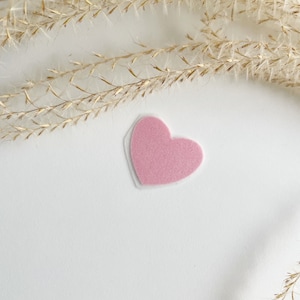 Könnte beinhalten: Ein pinkfarbenes Filzherz auf weißem Hintergrund mit getrocknetem beigefarbenem Gras im Hintergrund.