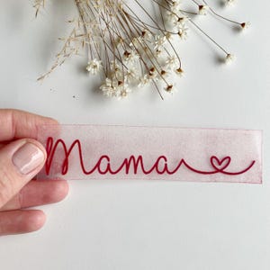 Könnte beinhalten: Ein transparentes Blatt mit dem Wort "Mama" in roter Kursivschrift und einem kleinen roten Herz am Ende.