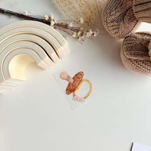 Könnte beinhalten: Aquarell-Illustration eines Babyschnullers mit braunem Sauger und gelbem Ring, verziert mit einer kleinen Schleife. Ein Holz-Regenbogenspielzeug und eine Strickmütze sind ebenfalls zu sehen, was auf ein Baby-Thema hindeutet.