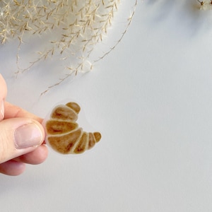 Könnte beinhalten: Ein transparenter Aufkleber mit einer braunen Croissant-Illustration.