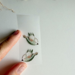 Könnte beinhalten: Zwei kleine, durchscheinende Aufkleber mit Aquarell-Illustrationen von fliegenden Vögeln. Die Vögel sind in gedeckten Grün- und Brauntönen gehalten, mit geschlossenen Augen und ausgebreiteten Flügeln dargestellt und vertikal auf einer Folie angeordnet.