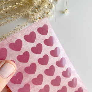 Könnte beinhalten: Eine transparente Vinylfolie mit einem rosa, gesprenkelten Hintergrund. Die Folie ist mit einem sich wiederholenden Muster aus rosa Herzen bedeckt.