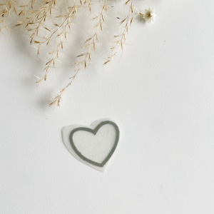 Könnte beinhalten: Ein herzförmiges Applikationsmotiv mit grauem Umriss und transparentem Zentrum. Die Applikation befindet sich auf einer weißen Oberfläche, mit getrocknetem Ziergras und einer kleinen weißen Blume im Hintergrund.