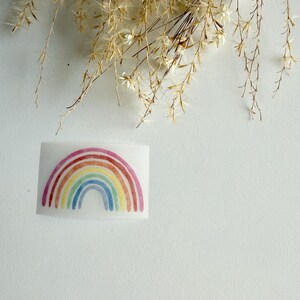 Könnte beinhalten: Ein rechteckiger Aufkleber mit einem Regenbogen im Aquarellstil, bestehend aus konzentrischen Bögen in den Farben Rot, Orange, Gelb, Grün, Blau und Violett. Der Regenbogen ist auf weißem Hintergrund gedruckt und zeigt eine weiche, texturierte Optik.