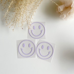 Könnte beinhalten: Drei violette Bügeletiketten mit Smiley-Gesichtern auf weißem Hintergrund.