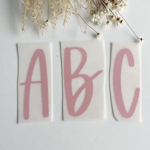 Könnte beinhalten: Drei rosa Vinyl-Aufkleber mit den Buchstaben A, B und C in handschriftlicher Form.