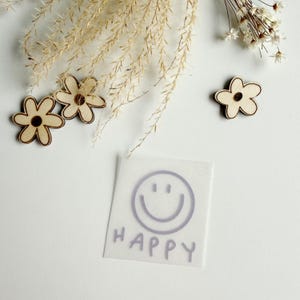 Könnte beinhalten: Weißer Hintergrund mit drei Holzblumen-Ausschnitten, getrockneten Pflanzen und einem kleinen Quadrat mit einem lila Smiley und dem Wort "HAPPY". Die Holzblumen haben eine hellbraune Farbe mit einem dunkleren braunen Zentrum.