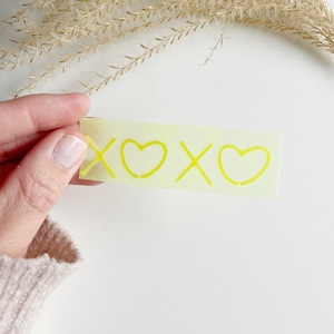 Könnte beinhalten: Ein rechteckiger, hellgelber Aufkleber mit dem Text "XOXO" in leuchtendem Gelb. Das Design zeigt zwei Herzen und zwei X. Der Aufkleber wird von einer Hand vor einem weißen Hintergrund gehalten.
