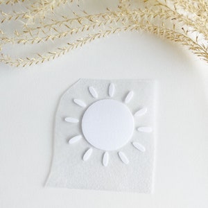 Könnte beinhalten: Ein weißes, sonnenförmiges Aufbügel-Transfermotiv mit einer soliden kreisförmigen Mitte, die von zwölf gleichmäßig verteilten ovalen Strahlen umgeben ist. Das Design befindet sich auf einer transparenten, rechteckigen Klebefolie.