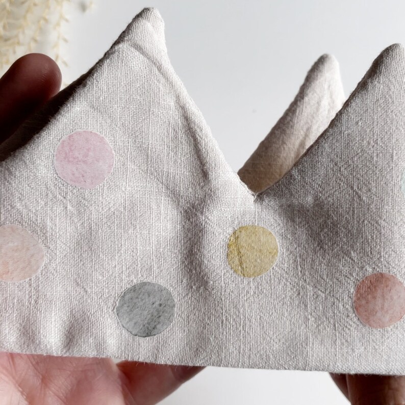 May include: A light gray fabric crown with a pointed top and a row of colorful polka dots. The dots are in shades of pink, yellow, and gray.