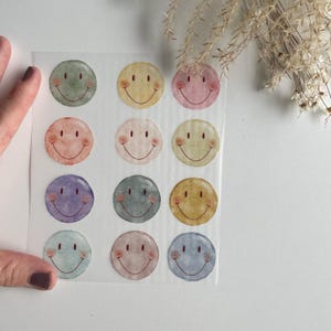 Könnte beinhalten: Eine Blatt mit bunten Smiley-Aufklebern mit aquarellartigen Designs. Die Aufkleber befinden sich auf einem transparenten Hintergrund und sind in einem Raster angeordnet.