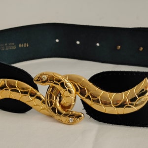 Op de afbeelding: Een zwarte riem met een goudkleurige metalen gesp in de vorm van twee in elkaar grijpende slangen met gestructureerde schubben en kleine heldere steentjes in de slangenkoppen. De binnenkant bevat de tekst 1098 CROUTE DE CUIR MADE IN FRANCE.