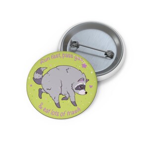 Op de afbeelding: Een ronde button met een speld aan de achterkant, voorzien van een lichtgroene achtergrond en een illustratie van een wasbeer. De tekst luidt, Run fast, pass gas, & eat lots of trash. Het ontwerp bevat kleine paarse sterretjes en hartjes.