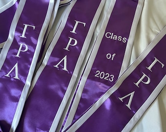 Może przedstawiać: Trzy fioletowe i białe szarfy absolwentów z haftem "Gamma Pi Lambda" i "Class of 2023" w kolorze białym.