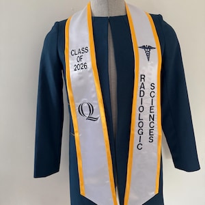 以下が含まれることがあります： 水色の卒業ガウンと白と金のサッシュ。サッシュには「CLASS OF 2026」と「RADIOLOGIC SCIENCES」の文字と医療シンボル、大きな「Q」のロゴが入っています。