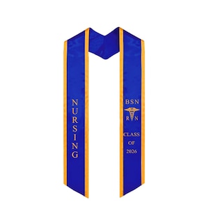 May include: A blue and gold graduation sash for nursing students. The sash has the word "NURSING" printed vertically in gold on one side and "BSN RN CLASS OF 2026" with a caduceus symbol on the other side.
