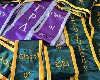 May include: A collection of green and purple graduation sashes with gold trim. The sashes have the text "Class of 2023" and "Radiology" embroidered on them.