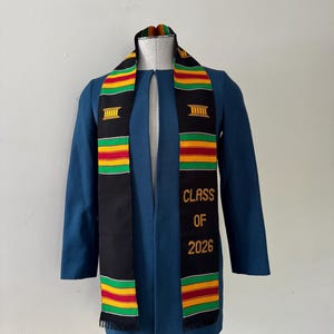 以下が含まれることがあります： 青緑色の卒業ガウンと、黒とカラフルなストール。ストールには「CLASS OF 2026」と金色の文字が書かれています。ストールには、緑、黄、赤のストライプ模様があります。