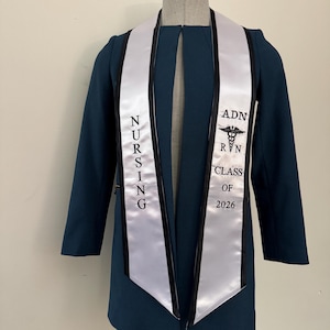 May include: A teal blazer with a white graduation sash. The sash has black trim and features the text "NURSING" vertically on one side and "ADN RN CLASS OF 2026" with a caduceus symbol on the other.