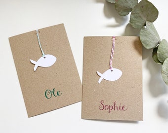 May include: Two brown cards with white fish shapes hanging from string. The card on the left says "Ole" in green lettering. The card on the right says "Sophie" in red lettering.