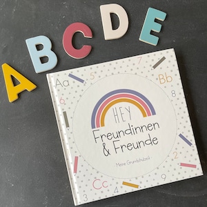 Könnte beinhalten: Ein farbenfrohes Buch mit dem Titel "HEY Freundinnen & Freunde - Meine Grundschulzeit" mit einem Regenbogen-Design und einem Kreis aus Zahlen und Buchstaben um den Regenbogen. Die Buchstaben A, B, C, D und E sind in verschiedenen Farben auf schwarzem Hintergrund dargestellt.