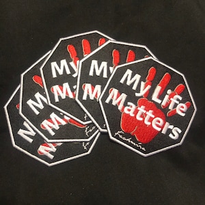 May include: Several black, octagonal patches with white embroidered edges. Each patch displays the text "My Life Matters" in white, with a red handprint design. The patches are arranged on a black background.