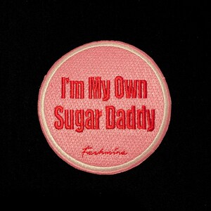 Pode incluir: Um patch redondo bordado em rosa com o texto "I'm My Own Sugar Daddy" em fio vermelho. O patch tem uma borda creme e a palavra "Fashionina" bordada na parte inferior.