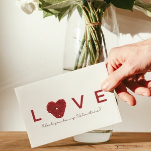 May include: A white card with the word "LOVE" in red letters and a red crocheted heart. The text below the word "LOVE" reads "Will you be my Valentine?"