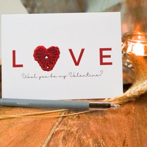 May include: A white card with the word "LOVE" in red letters. A red crocheted heart replaces the "O". The text "Wool you be my Valentine?" is written in cursive below the word "LOVE".