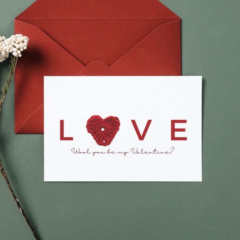 May include: A white card with the word "LOVE" in red letters and a red crocheted heart in the centre. The text below the word "LOVE" reads "Wool you be my Valentine?"