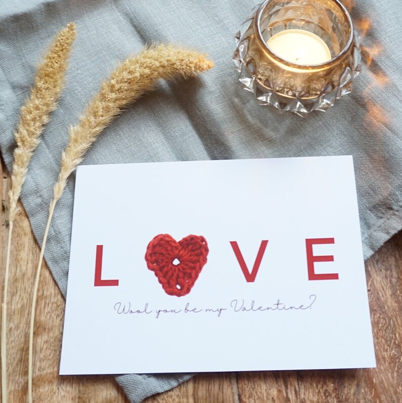 May include: A white card with the word "LOVE" in red letters. A red crocheted heart is in the middle of the word. The text "Wool you be my Valentine?" is written below the word "LOVE".