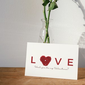 May include: A white card with the word "LOVE" in red letters. A red crocheted heart is in the middle of the word. The text "Wool you be my Valentine?" is written below the word "LOVE".