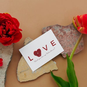 May include: A white card with the word "LOVE" in red lettering and a red heart with a white centre. The card says "Will you be my Valentine?"  A red flower and a rock are in the background.