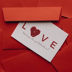 May include: A white card with the word "LOVE" in large letters and a red crocheted heart. The text on the card reads "Wool you be my Valentine?" The card is lying on top of several red envelopes.
