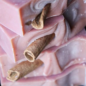 Könnte beinhalten: Ein Stapel rechteckiger Seifenstücke in Mauve, jedes mit einer glatten, gewirbelten Textur und einem einzelnen, ganzen braunen Zimtstangenstück, das aus der Mitte der Vorderseite jedes einzelnen Seifenstücks herausragt.