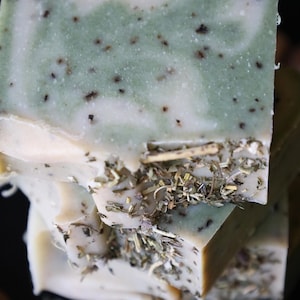 May include: A stack of handmade soap bars. The top bar has a light green and white marbled design with dark specks and dried herbs. The soap is stacked on top of other bars, with a dark background.