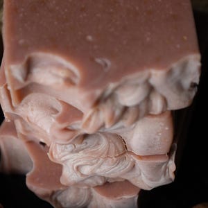 Könnte beinhalten: Nahaufnahme von handgemachten Seifenstücken. Die Seife hat einen hellrosa Farbton mit einer strukturierten Oberfläche und sichtbaren Flecken. Die Kanten der Seife haben ein dekoratives, wirbelndes Design. Die Seife ist gestapelt, wobei die unteren Stücke ein detailliertes, geschichtetes Muster aufweisen.