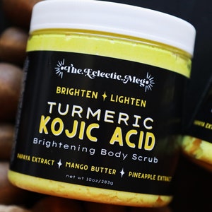May include: A yellow jar of turmeric and kojic acid brightening body scrub with a black label that reads "The Eclectic Meg", "Brighten + Lighten", "Turmeric Kojic Acid", "Brightening Body Scrub", "Papaya Extract", "Mango Butter", "Pineapple Extract", and "net wt 10oz/283g".