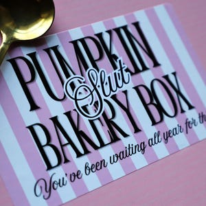 May include: A rectangular card with a light pink and white vertical striped background. The text reads PUMPKIN SLUT BAKERY BOX in black, with the phrase You've been waiting all year for this printed below. A gold-toned metal spoon rests on the left.