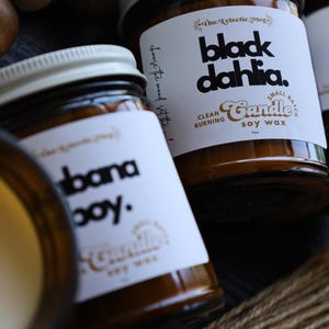 May include: Three amber glass jars containing soy wax candles with white labels. The labels feature black text reading black dahlia, cabana boy, and cafe rosa, along with the text clean burning soy wax and the eclectic meg branding.