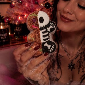 May include: A dual-sided bath bomb shaped like a gingerbread person, featuring a brown textured side and a white skeleton design on the other side. The bath bomb is held up, showcasing its split aesthetic with a detailed skeletal print.