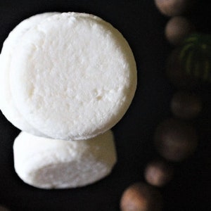 May include: Two white round solid shampoo bars stacked on top of each other. The bars are on a black surface.