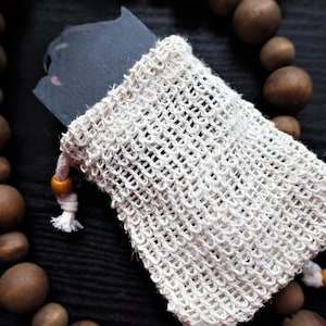 May include: A black bar of soap in a white mesh bag with a wooden bead drawstring. The bag is on a dark wooden surface with brown wooden beads.