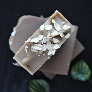 May include: A stack of two rectangular bars of soap with a light brown color. The top bar is covered in oat flakes.